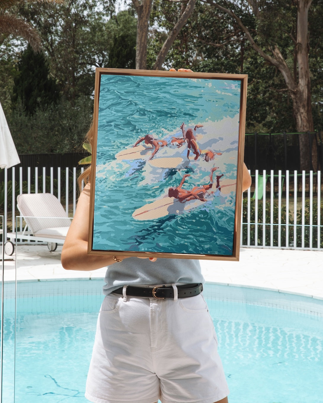 Paint Like Frida's Chaos Big Framed Canvas in a custom-made floating frame, featuring five surfers diving into turquoise waves — held poolside by a woman in white shorts, with sunny summer vibes all around.